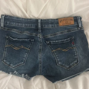 Blå jeansshorts från Replay - Snygga blå jeansshorts från Replay med slitna detaljer och råa kanter. Klassisk femficksmodell med dragkedja och knapp framtill. Bakfickorna har dekorativa sömmar och Replay-logga. Perfekta för en avslappnad stil under varma dagar.