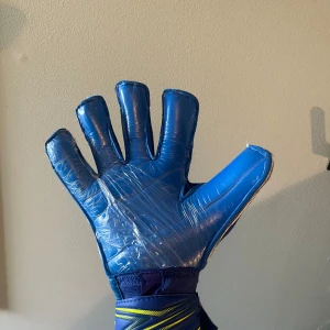 RG Bacan målvaktshandskar blå/gul - Säljer ett par feta RG Bacan målvaktshandskar i blått och gult med coolt grafiskt mönster. Handflatan är helblå och ovansidan har snygga detaljer i gult och blått. Breda kardborreband för extra stöd runt handleden. Perfekta för dig som lirar fotboll och vill sticka ut på planen. Helt nya!!Storlek 10 och pris 500kr!