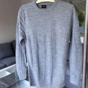 Grå stickad tröja  - Säljer en grå stickad tröja från Hazar-De. Tröjan har ett snyggt kabelstickat mönster upptill och vertikala ränder nedtill. Rund halsringning och ribbade muddar vid ärmslut och nederkant. Oanvänd.