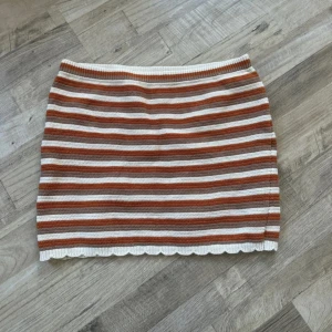 Randig stickad kortkjol i brunt och vitt - Superfin stickad kortkjol med horisontella ränder i brunt och vitt. Kjolen har en rak passform och söt vågig kant längst ner. Perfekt för dig som gillar retrovibbar och vill ha något bekvämt men ändå snyggt.