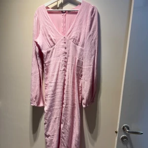 Ljusrosa långklänning från Nelly - Säljer denna superfina ljusrosa långklänning med v-ringning, långa puffärmar och knappar framtill. Klänningen har en snygg slits och är gjord i ett lätt, strukturerat tyg som känns luftigt. Perfekt för sommarens speciella tillfällen! Den är använd 1 gång och har tyvärr inte så fått några fler tillfällen att ha denna skönhet på. Nypris 700kr