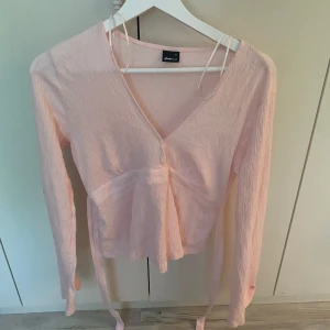 Ljusrosa omlottopp från Gina Tricot - Supersöt ljusrosa omlottopp från Gina Tricot med långa ärmar och v-ringning. Toppen har knytband i midjan som ger en snygg siluett och är gjord i ett lätt, krinklat tyg. Toppen är sydd i v-ringningen då den var för stor för mig, men går enkelt att sprätta upp!🫰