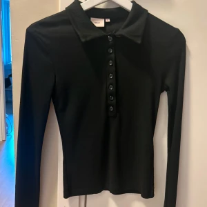 Svart ribbad pikétröja från Nelly - Svart långärmad pikétröja från Nelly med ribbad struktur och krage. Tröjan har knappar framtill . Perfekt till jeans eller kjol för en clean look. Aldrig använd, köpt för 199kr och säljs för 50kr