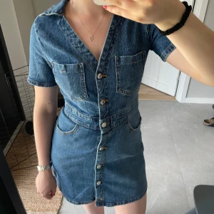 Jeansklänning med knappar och fickor - Snygg jeansklänning i blå denim med kort ärm, v-ringning och knappar hela vägen framtill. Helt oanvänd! 