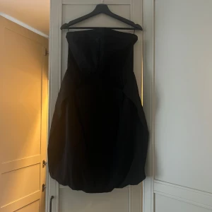 Svart axelbandslös kortklänning - Snygg svart axelbandslös kortklänning med ballongformad kjol. Klänningen har en stilren och elegant design som ger en cool siluett. Perfekt för dig som vill sticka ut med en klassisk men modern look.