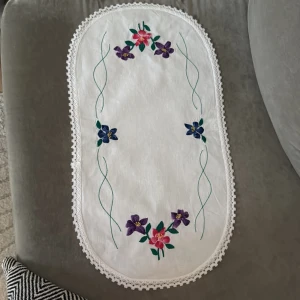 Broderad duk - Bilderna visar en vackert broderad duk med blommotiv och spetskant. Duken har lila, blå och rosa blommor samt gröna blad, och är perfekt för att ge ditt rum en vintage och personlig känsla. Passar dig som gillar unika detaljer och handarbete.