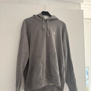 Grå hoodie från Polo Ralph Lauren - Säljer en grå hoodie från Polo Ralph Lauren med klassisk huva och snörning. Tröjan har en liten vit logga på bröstet och en stor känguruficka framtill.  Passar M/L och är väldigt fin. Nypris-2.4