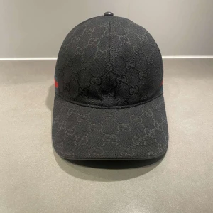 Svart keps från Gucci med GG-mönster - Svart keps från Gucci (Aldrig använt) med diskret GG-monogram över hela ytan och klassiska gröna och röda ränder på sidan. Justerbar rem baktill och insida med Gucci-etikett. Snygg och stilren accessoar för dig som gillar exklusiva märken.