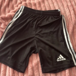 Svarta Adidas shorts med vita ränder - Svarta sportiga Adidas shorts med klassiska vita ränder längs sidorna och Adidas-logga framtill. Elastisk midja för skön passform. Perfekta för träning eller chill. Lätta och svala i syntetmaterial.