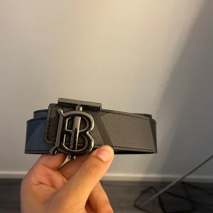 Burberry svart rutigt skärp med B-spänne - Snyggt svart bälte från Burberry med klassiskt rutigt mönster i grå toner. Bältet har ett stilrent metallspänne med den ikoniska B-loggan framtill. Tillverkat i exklusivt skinn med en lyxig känsla och modern design.