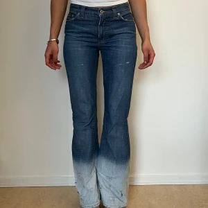 Blå bootcut jeans från Dolce & Gabbana - Säljer ett par blå bootcut jeans från Dolce & Gabbana med snygg dip dye-effekt som går från mörkblå upptill till ljusblå nertill. Jeansen har slitna detaljer vid bensluten och klassisk femficksdesign. Perfekta för dig som vill sticka ut! 