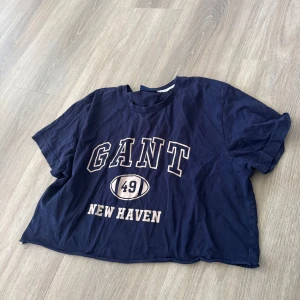 Mörkblå croppad t-shirt från GANT - Mörkblå croppad t-shirt från GANT med tryck 'GANT 49 NEW HAVEN' framtill. Klassisk rund halsringning och korta ärmar. Perfekt för en avslappnad och sportig stil.