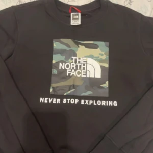 Svart sweatshirt från The North Face med camo-tryck - Säljer en svart sweatshirt från The North Face med ett stort kamouflage-tryck och logga på bröstet. Texten 'Never Stop Exploring' under trycket. Klassisk rund hals och långa ärmar. Perfekt för en avslappnad stil.