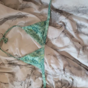 Grön paisleymönstrad bikiniöverdel från Gina Tricot - Säljer en grön bikiniöverdel från Gina Tricot med paisleymönster och tunna knytband. Triangelformad modell med mjukt tyg och dekorativ kant. Perfekt för sommardagar vid stranden eller poolen.