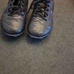 Svarta Nike fotbollsskor med blå detaljer. - Säljer ett par svarta Nike fotbollsskor med blå detaljer och vit Nike-logga på sidan. Skorna har snörning och är tillverkade i syntetmaterial. Perfekta för dig som vill ha stilrena och funktionella skor på planen.