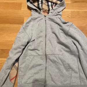 Burberry hoodie - Säljer en stilren grå hoodie från Burberry med dragkedja och fickor framtill. Insidan av huvan och ärmsluten har det ikoniska Burberry-rutiga fodret i beige, svart, rött och vitt. Perfekt för dig som gillar klassisk design med en twist. Hoodien har rutigt mönster på armbågarna. DET ÄR STORLEK 164CM MEN PASSAR XS SKULLE JAG SÄGA…