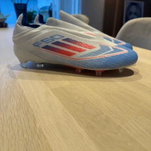 Adidas F50  - Säljer ett par Adidas F50 fotbollsskor i vitt och blått med röda detaljer. Skorna har en strömlinjeformad design utan snörning och mönstrad ovandel. Perfekta för dig som vill ha lätta och snabba skor på planen. Bara testade en gång, säljer för att de va för stora 