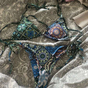 Flerfärgad bikini med knyt och mönster - Säljer en snygg bikini med färgglada geometriska och blommiga mönster i blått, grönt och inslag av rött. Både överdel och underdel har knytband i sidorna och nacken. Perfekt för sommaren och strandhäng!