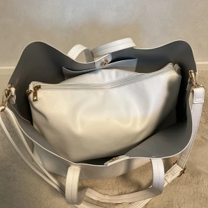 Vit shoppingväska och axelväska - Säljer en stor vit shoppingväska med matchande, oanvänd, ”innerväska” i syntetmaterial. Shoppingväskan har två handtag och enkel design, medan den minde fungerar mer som en necessär med dragkedja. Finns även ett justerbart axelband som fungerar att sätta på både den större och mindre väskan. Shoppingväskan har många skavanker och ser väldigt använd ut men fungerar bra. Den lilla är helt oanvänd och lika så axelremmen. 