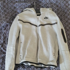 Ljusgrå Nike Tech Fleece  - Nike hoodiejacka i ljusgrått med svarta detaljer och dragkedja framtill. Jackan har huva, lång ärm med extra ficka med dragkedja på ena ärmen och svarta ränder över bröstet. Perfekt för en sportig look.