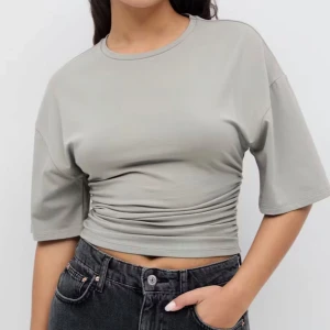 Beige croppad t-shirt med rynkade detaljer - Trendig beige croppad t-shirt med korta ärmar och rund halsringning. Snygga rynkade detaljer i midjan ger en cool och modern look. Perfekt till jeans eller kjol för en avslappnad stil.