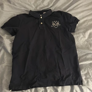 Svart pikétröja från GANT - Säljer en mörkblå pikétröja från GANT med klassisk krage och två knappar framtill. På bröstet finns ett broderat GANT-märke med tennismotiv. Perfekt för en sportig och stilren look.