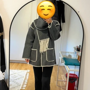 Mörkgrå kappa med dekorativa sömmar och matchande halsduk  -  Mörkgrå kappa med vita dekorativa sömmar och stora fickor. Ser helt ny ut. Kappan stängs med knappar. Kappan har en medföljande halsduk med fransar. Rak passform och snygga detaljer längs kanterna. Stilrent och modernt✨