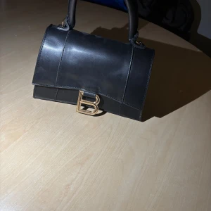 Svart handväska  - Säljer en stilren svart handväska  med ett stort B-spänne i metall framtill. Väskan har ett klassiskt, fyrkantigt format och bärhandtag upptill. Perfekt för dig som gillar tidlös design och vill ha en ikonisk accessoar.