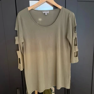 Olivgrön topp med unika ärmdetaljer - Snygg olivgrön topp med trekvartsärm och öppna detaljer längs ärmarna. Rund halsringning och rak passform. Perfekt för dig som vill ha något annorlunda och stilrent.