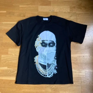 Svart t-shirt med konstnärligt tryck - Svart t-shirt med ett stort konstnärligt tryck av ett ansikte med smycken och mask på framsidan. T-shirten har rund hals och korta ärmar. Perfekt för dig som vill sticka ut med din stil.