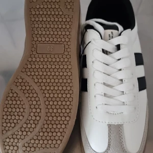 Vita sneakers med svarta ränder - Säljer ett par klassiska vita sneakers från lager 157 med svarta ränder på sidorna. Skorna har vit snörning, beige sula och detaljer i mocka framtill. Perfekta för en sportig och stilren look.               Storlek 39 och är oanvänd. 