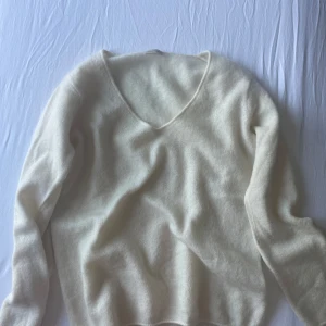Vit ulltröja från By Malene Birger - Mjuk och fluffig vit tröja från By Malene Birger i ullblandning. Tröjan har v-ringning och långa ärmar, perfekt för kyligare dagar. Enkel och stilren design som passar till mycket.