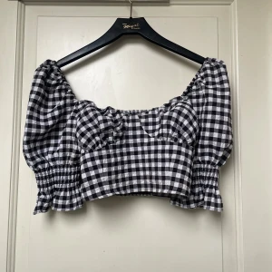 Svartvit rutig croppad blus - Trendig croppad blus med svartvitt rutigt mönster och puffiga korta ärmar. Smockad baktill och vid ärmslut för extra volym. Perfekt för en stilren och lekfull look.