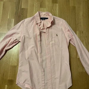 Ljusrosa skjorta från Ralph Lauren - Säljer en klassisk ljusrosa skjorta från Ralph Lauren i slim fit-modell. Skjortan har långa ärmar, knappar framtill och den ikoniska broderade loggan på bröstet. Perfekt för dig som gillar stilrena plagg med snygg passform.