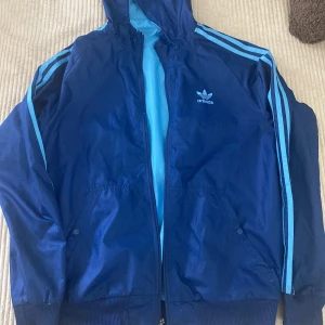 Blå vindjacka från Adidas - Säljer en blå vindjacka från Adidas med huva och klassiska ränder på ärmarna. Jackan är vändbar med en mörkblå och en ljusblå sida, båda med dragkedja och fickor. Adidas-logga på bröstet. Perfekt för dig som gillar sportig stil.