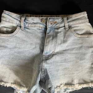 Ljusblå jeansshorts från Primark - Ljusblå jeansshorts från Primark med fransig kant nertill och klassisk femficksdesign. Shortsen har bälteshällor och knappstängning framtill. Perfekta för varma dagar och enkel att matcha med olika toppar.