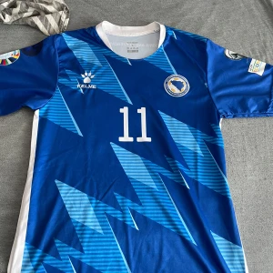 Blå Bosnien och Hercegovina fotbollströja med nummer 11 - Säljer en blå fotbollströja från Bosnien och Hercegovina med nummer 11 och namnet Džeko på ryggen. Tröjan har vita detaljer, korta ärmar och ett mönster av ljusblå blixtar. Perfekt för fans av landslaget.