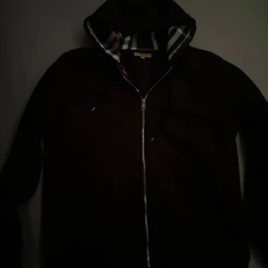 Svart hoodiejacka från Burberry - Säljer en svart hoodiejacka från Burberry med klassiskt rutigt foder i huvan och insidan. Jackan har dragkedja framtill och broderad logga på bröstet. Perfekt för dig som gillar stilrena detaljer och ikoniskt mönster. Passar L perfekt 🤩
