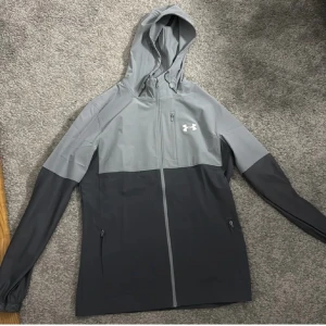 Grå vindjacka med huva från Under Armour - Säljer en snygg tvåfärgad grå vindjacka från Under Armour med huva och dragkedja framtill. Jackan har två sidofickor med dragkedja och en sportig look, perfekt för utomhusaktiviteter.