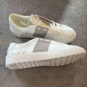 Valentino open  - Säljer ett par snygga vita sneakers från Valentino Garavani med bred grå panel på sidan och vita snören. Skorna har rund tå, platt sula och diskreta nitar på sidan av sulan. Perfekta för dig som gillar stilrena och exklusiva sneakers.
