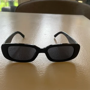 Svarta rektangulära solglasögon - Snygga svarta solglasögon med rektangulära bågar och mörka glas. Perfekt accessoar för en stilren look. Bågarna är i plast och har en klassisk, minimalistisk design.