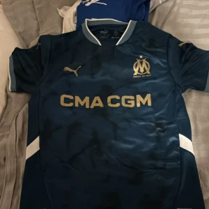 Marseille fotbollströja Puma - Mörkblå Marseille fotbollströja från Puma med guldiga detaljer och logga. Kortärmad modell med vit krage och vita inslag på ärmarna. Tryckt sponsortext på bröstet. Helt ny 