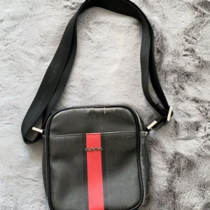 Svart väska Valentino - Snygg svart axelväska från Valentino med en röd vertikal rand på framsidan. Väskan har justerbart axelband, dragkedja både fram och upptill samt metall-detaljer med logga. Detaljer som liknar ”rivna jeans” stilen, unik stil & köpt från Zalando