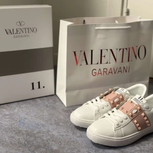 Valentino Garavani sneakers med nitar - Säljer ett par vita sneakers från Valentino Garavani med breda ljusrosa band och ikoniska nitar i guld längs sidorna och bak på skon. Skorna har rund tå, platt sula och klassisk snörning. Perfekta för dig som gillar lyxiga detaljer och stilren design.
