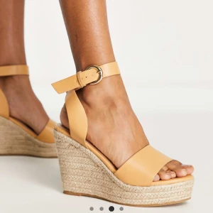 Beige kilklackade sandaler från Glamorous - Säljer ett par snygga beige sandaler från Glamorous asos med hög kilklack i rep och breda remmar. Skorna har öppen tå och justerbart spänne vid ankeln. Perfekta för sommaren och ger en trendig look till din outfit.