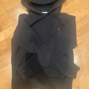 Svart hoodie från Polo Ralph Lauren - Svart hoodie från Polo Ralph Lauren med klassisk röd broderad logga på bröstet. Tröjan har huva och en stor magficka framtill. Perfekt för en avslappnad och stilren look.