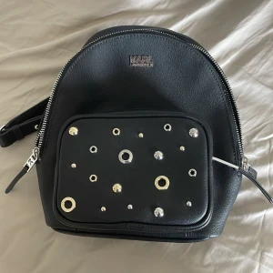 Svart ryggsäck med nitar från Karl Lagerfeld - Säljer en svart äkta mini ryggsäck från Karl Lagerfeld med silvriga nitar och detaljer på ytterfacket. Väskan har justerbara axelremmar, dragkedja och ett mönstrat innerfoder. Den har två fack, ett större och ett mindre därfram. Väskan är liten i sig men rymmer en hel del ändå. Perfekt för dig som vill ha en cool och stilren accessoar. Pris kan diskuteras, men kommer inte sänka alldeles för mycket eftersom att den är helt sprillans ny.