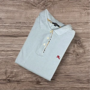 Ljusblå pikétröja från Burberry - Säljer en ljusblå pikétröja från Burberry med klassisk krage och knäppning framtill. Tröjan har korta ärmar och en liten röd broderad logga på bröstet. Typisk Burberry-detalj vid knappslån. Perfekt för en stilren look.