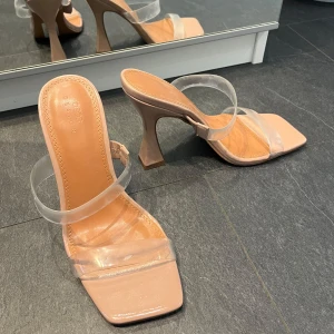 Beiga klackskor med transparenta remmar - Säljer ett par snygga beiga klackskor med fyrkantig tå och två breda transparenta remmar. Skorna har hög klack och en stilren design som passar perfekt till sommarens outfits.