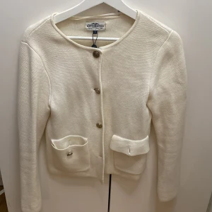 Vit cardigan jacka från Kappahl  - Säljer en stilren vit cardigan jacka från Gina Tricot med rund halsringning och dekorativa knappar framtill. Jackan har två fickor med lock och är tillverkad i ett mjukt stickat material. Perfekt för lager-på-lager och enkel att matcha.